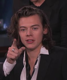 a man with long hair wearing a black suit and white shirt pointing to the side