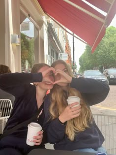 two women sitting at an outdoor table with coffee cups in front of them, making a heart shape