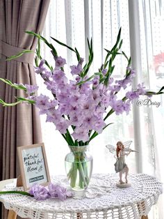 a glass vase with purple flowers on a table next to a small fairy figurine