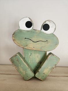 a green frog sitting on top of a wooden table next to a white wall with black eyes