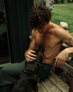 a shirtless man sitting on a wooden bench with his black dog next to him