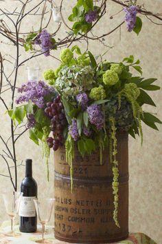 a wooden barrel filled with lots of purple flowers and greenery next to a bottle of wine