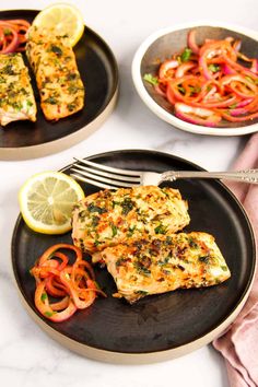 two black plates with grilled salmon and vegetables on them, one plate has lemon wedges