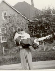 an old black and white photo of a man holding a woman on his back in front of a house