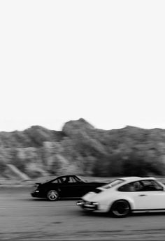 black and white photograph of two cars driving down the road with mountains in the background