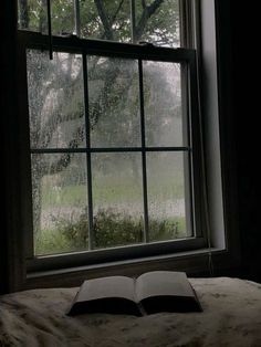 an open book sitting on top of a bed in front of a window covered in rain