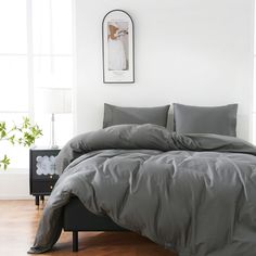 a bed with grey sheets and pillows in a white room next to a wooden floor