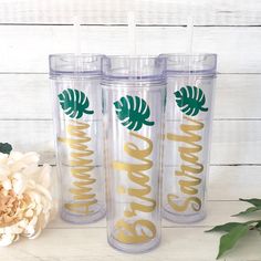 three personalized tumblers with gold lettering and palm leaves on them, sitting next to a flower