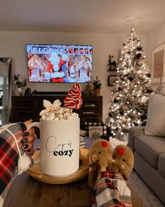 a person holding a mug with marshmallows and candy in front of a christmas tree