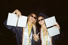 two young women in graduation gowns holding diplomas and pointing to something off camera