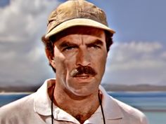 a man with a mustache wearing a hat and white shirt is standing in front of the ocean