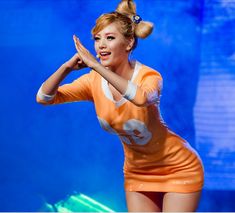 a woman in an orange dress on stage with her hands up to the side and one hand behind her head