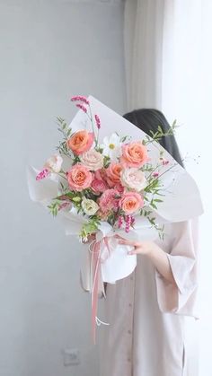 a woman holding a bouquet of pink and white flowers in front of her face while standing next to a window