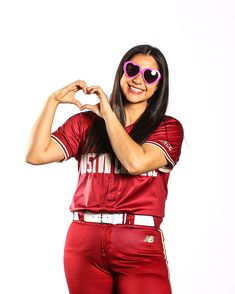 a woman in a baseball uniform making a heart shape with her hands while standing against a white background