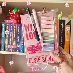 a person holding up a planner and pen in front of a book shelf with books on it