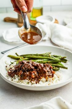 a person is spooning sauce over some food on a white plate with rice and asparagus