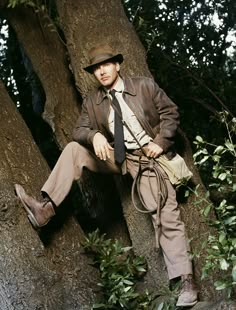 a man sitting on top of a tree wearing a hat and tie with his hands in his pockets