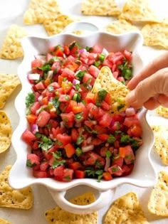 a person dipping salsa into a white bowl with tortilla chips on the side