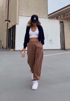 a woman walking across a parking lot holding a coffee cup and wearing a baseball cap