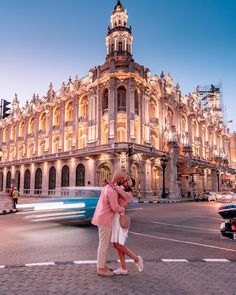 two people walking across a street in front of a large building with a clock tower