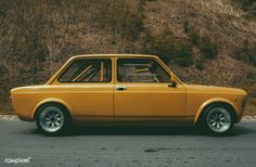 an old yellow car is parked on the side of the road in front of a hill