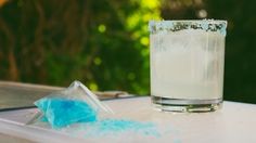 a glass filled with blue substance next to a bottle of liquid on top of a table