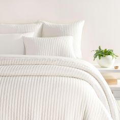 a bed with white comforter and pillows on it next to a potted plant