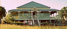 three people standing in front of a green house with tall grass and palm trees behind them