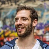 a man with a beard smiles in front of graffiti covered walls and trees, wearing a blue hoodie