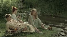 three women sitting on the ground next to a river and some grass in front of them