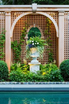 an outdoor fountain surrounded by greenery and flowers in front of a pergolated wall