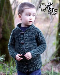 a young boy standing in the woods wearing a green knitted cardigan and jeans