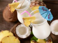 two glasses filled with pineapple milkshakes next to coconuts and an umbrella