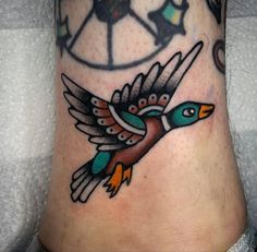 a colorful bird tattoo on the ankle with compasss in the background and birds flying around it