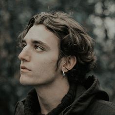 a young man with curly hair wearing a black jacket and earrings looking off into the distance