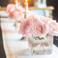 pink roses are in a square vase on a table with white linens and candles