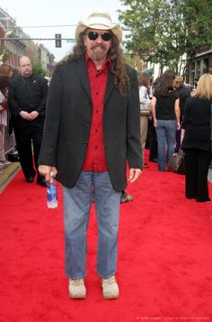 a man with long hair wearing a hat and sunglasses standing on a red carpeted area