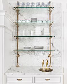 a white kitchen with marble counter tops and gold faucet, glass shelves above the sink