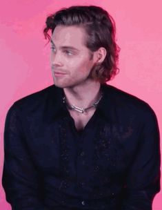 a man with long hair sitting down in front of a pink background wearing a black shirt