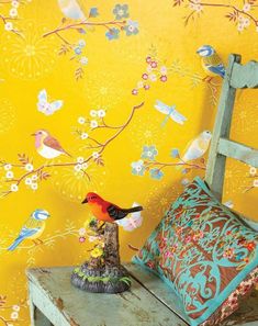 a small bird sitting on top of a wooden bench next to a yellow wall with flowers