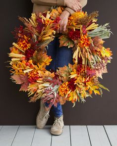 a woman is holding a wreath made out of fake leaves and autumn flowers on a wooden floor