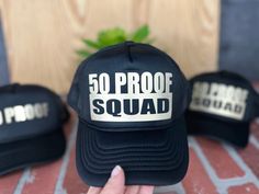 a person holding up a black hat with 50 proof squad written on the front and back