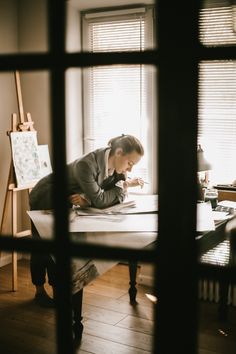 a woman sitting at a table writing on a piece of paper in front of a window