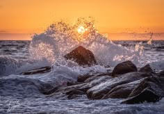 the sun is setting over the ocean with waves crashing on rocks in front of it