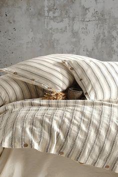 an unmade bed with striped sheets and pillows in front of a concrete wall,