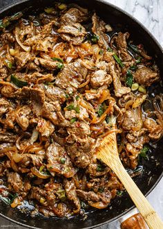 a skillet filled with meat and vegetables on top of a marble counter next to a wooden spatula