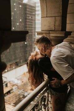 a man kissing a woman on the cheek while standing next to a railing with buildings in the background