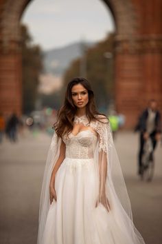 a woman in a white wedding dress and veil standing on the street with an archway behind her