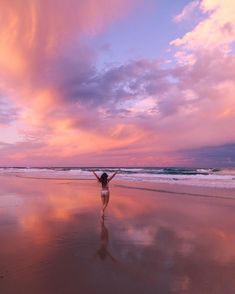 a woman standing on top of a sandy beach under a pink and blue sky with clouds