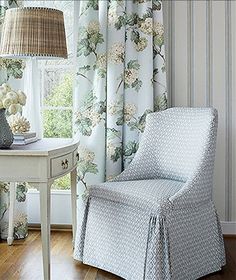 a white chair sitting in front of a window next to a table with a lamp on top of it